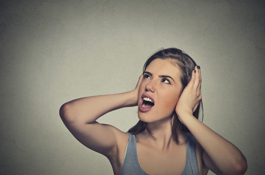 Closeup portrait young unhappy stressed woman covering her ears looking up stop making loud noise it's giving me headache isolated on gray wall background. Negative emotion face expression feeling
