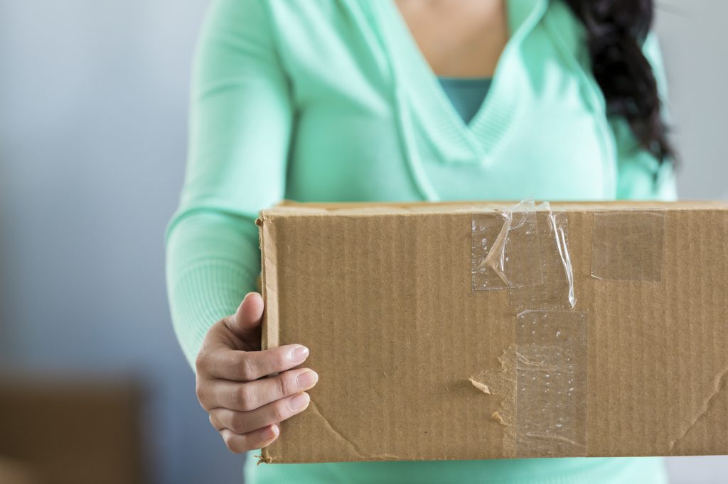 Woman is holding a packed moving box. She is preparing to move or unpack the box. The is wearing a light green blouse. focus in on the cardboard box. The box is uses with tape on it.