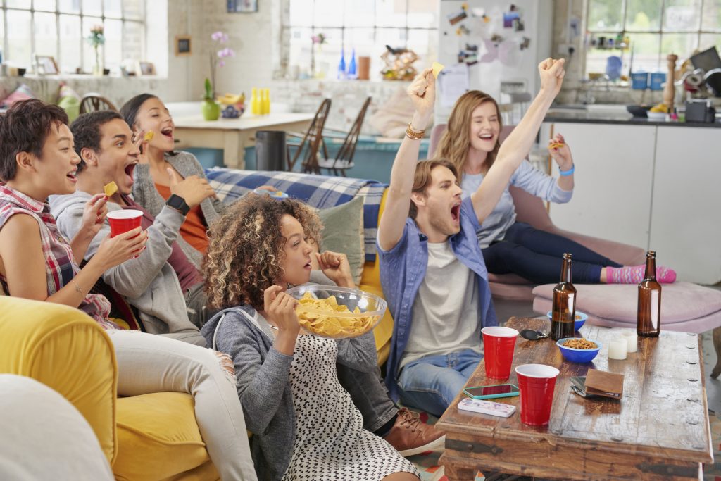 Diverse mix of friends sports fans watching winning football match on TV at home Celebrating winning goal huddled on couch shouting excited sharing snacks drinking beer